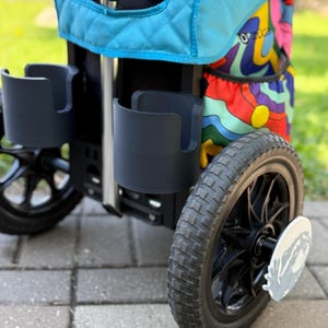 Puede incluir: Un cochecito negro y turquesa con dos portavasos y un compartimento de almacenamiento con estampado colorido. El cochecito tiene ruedas negras con una placa decorativa plateada en la rueda. El cochecito est&aacute; sobre una superficie de ladrillo.