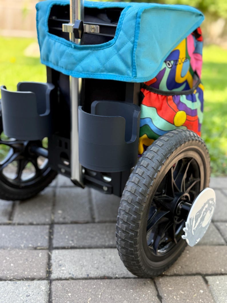 Puede incluir: Un cochecito negro y turquesa con dos portavasos y un compartimento de almacenamiento con estampado colorido. El cochecito tiene ruedas negras con una placa decorativa plateada en la rueda. El cochecito est&aacute; sobre una superficie de ladrillo.