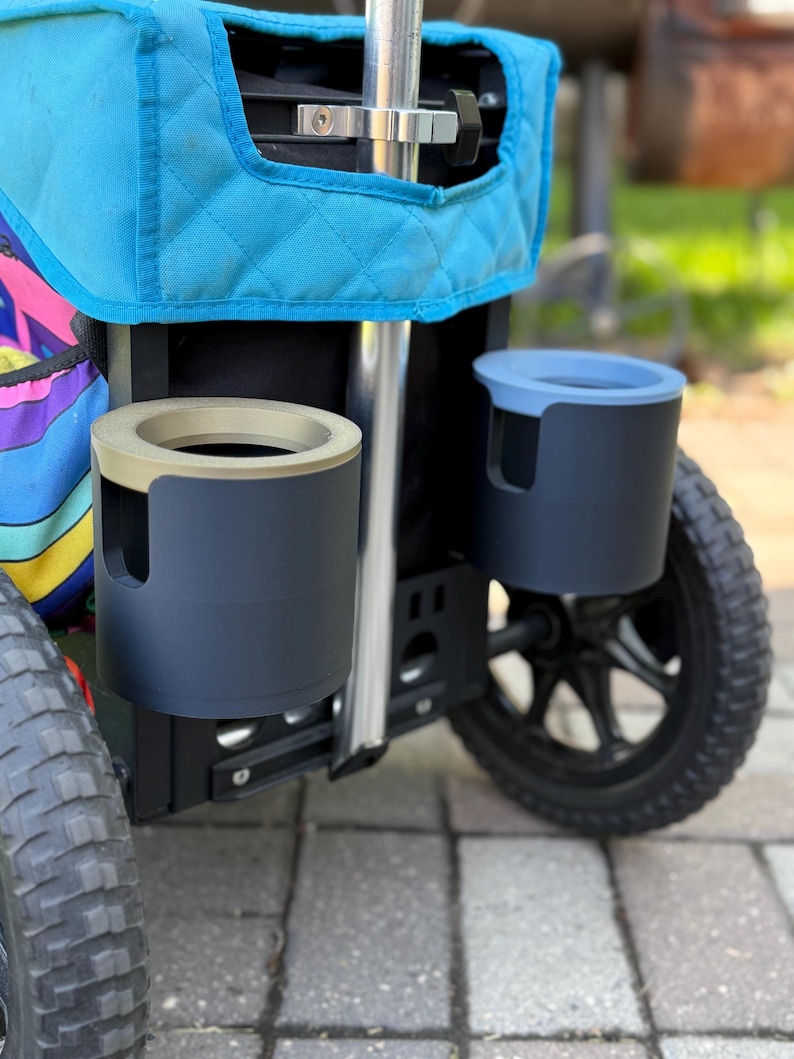 Puede incluir: Primer plano de un carrito utilitario negro con dos portavasos. Un portavasos es gris oscuro con un borde beige, y el otro es gris oscuro con un borde azul claro. El carrito tiene un &aacute;rea de almacenamiento acolchada azul y ruedas negras.