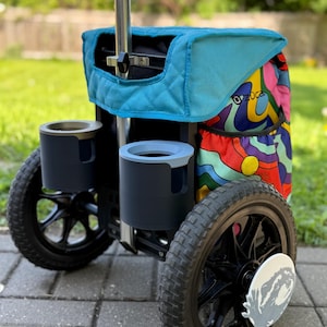 Puede incluir: Un carrito de disc golf Zuca con una parte superior acolchada de color turquesa y un lateral estampado colorido. El carrito tiene dos portavasos, ruedas negras y un marco plateado. Las ruedas tienen un di&aacute;metro de aproximadamente 25 cm.