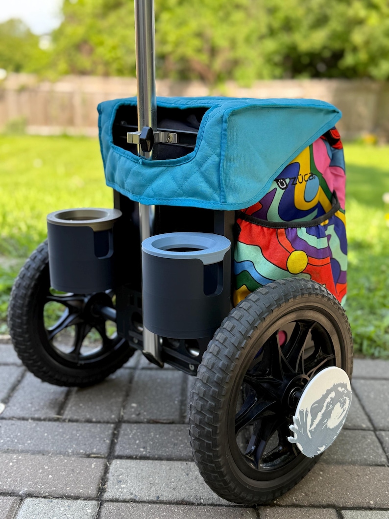 Puede incluir: Un carrito de disc golf Zuca con una parte superior acolchada de color turquesa y un lateral estampado colorido. El carrito tiene dos portavasos, ruedas negras y un marco plateado. Las ruedas tienen un di&aacute;metro de aproximadamente 25 cm.