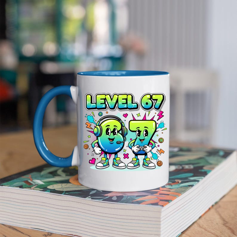 May include: White ceramic mug with a blue interior and handle. The mug features the text "LEVEL 67" above cartoon figures of the numbers 6 and 7, both with faces and holding game controllers. The mug is sitting on a book.