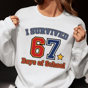 May include: White sweatshirt with the text "I Survived 67 Days of School". The number 6 is red with a yellow outline, and the number 7 is blue. The text "Days of School" is in orange.