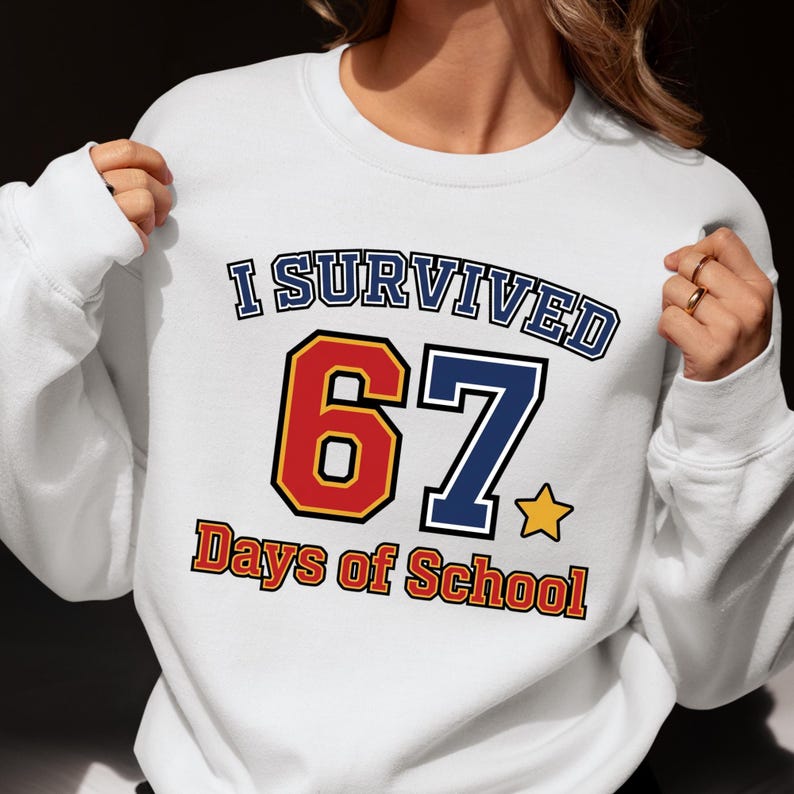 May include: White sweatshirt with the text "I Survived 67 Days of School". The number 6 is red with a yellow outline, and the number 7 is blue. The text "Days of School" is in orange.