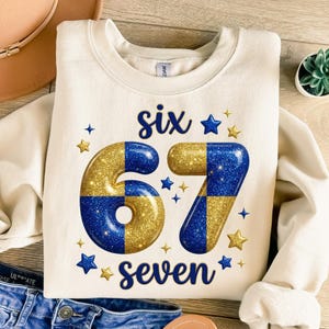 May include: Cream-coloured sweatshirt featuring the words "six seven" and the numbers 67 in a gold and blue chequered pattern. Blue and gold stars are scattered around the numbers and words.