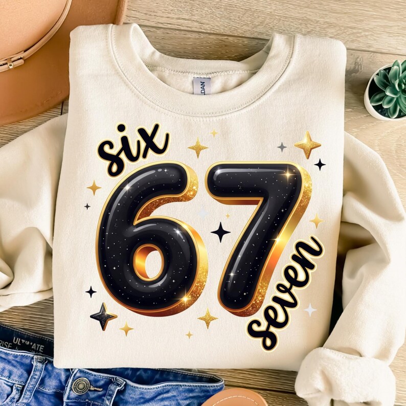 May include: Cream-coloured sweatshirt with the text "six 67 seven" in black and gold. The numbers are large and outlined in gold, with a starry design. The sweatshirt is folded, with a hat and jeans visible.