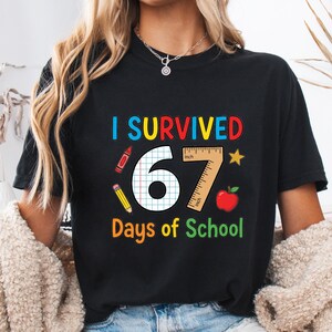 May include: Black t-shirt with the text "I Survived 67 Days of School" in colourful letters. The design includes a ruler, apple, star, and school supplies. The shirt is a crew neck.