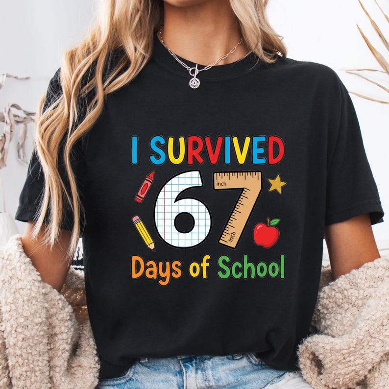 May include: Black t-shirt with the text "I Survived 67 Days of School" in colourful letters. The design includes a ruler, apple, star, and school supplies. The shirt is a crew neck.