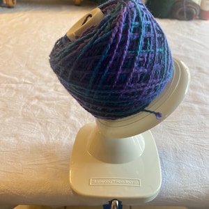 May include: A white yarn holder with a blue and purple yarn ball on top. The yarn holder has a clamp to attach it to a table or other surface. The yarn holder is labeled "Stanwood Needlecraft".