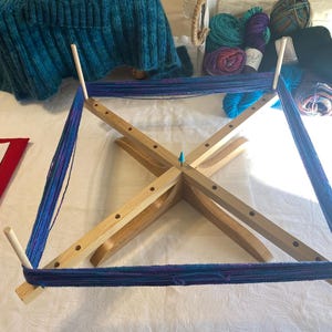 May include: A wooden yarn swift with four arms and a central post. The swift is holding a skein of blue and purple yarn. The yarn is wound around the arms of the swift and is being held in place by the central post.