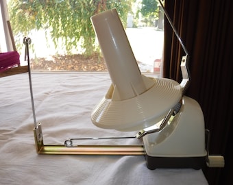 May include: A cream-colored yarn winder with a metal frame and a dark brown base. The winder is designed to hold yarn and create neat yarn cakes. A wooden yarn swift is visible in the background, holding a skein of pink yarn.