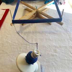 Może przedstawiać: Drewniany nawijacz do włóczki z niebieską i fioletową włóczką owiniętą wokół niego. Nawijacz do włóczki stoi na białym stole z białym nawijaczem do kłębków włóczki z niebieskim i fioletowym kłębkiem włóczki na nim.