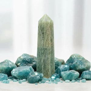 Könnte beinhalten: Ein hoher, hellgrüner Kristallobelisk steht inmitten verschiedener Größen von blau-grünen Rohkristallen. Der Obelisk ist der Mittelpunkt, umgeben von den grob geschnittenen Steinen. Die Kristalle variieren in Größe und Form und erzeugen eine natürliche, erdige Ästhetik.