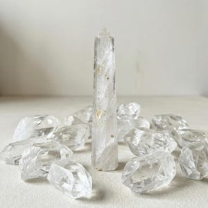 Könnte beinhalten: Eine Ansammlung klarer Quarz-Kristalle. Ein hoher, polierter Quarzpunkt steht aufrecht, umgeben von grob geschnittenen Quarzstücken. Die Kristalle sind durchscheinend mit feinen Goldflocken, vor einem neutralen Hintergrund.