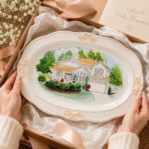 Könnte beinhalten: Eine ovale weiße Keramikplatte mit einer handgemalten Aquarellillustration eines zweistöckigen Hauses mit Garage, umgeben von grünen Bäumen und Pflanzen. Die Platte hat einen Perlenrand und dekorative erhabene Verzierungen.