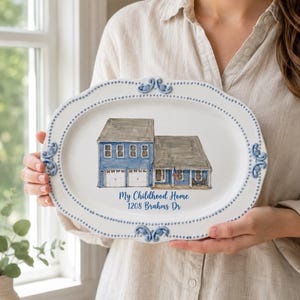 Op de afbeelding: Een ovale witte keramische serveerschaal met een kralenrand en blauwe decoratieve accenten. Het midden bevat een aquarelillustratie van een blauw huis met twee verdiepingen, een garage en de tekst "My Childhood Home 1208 Brahms Dr" in blauw.