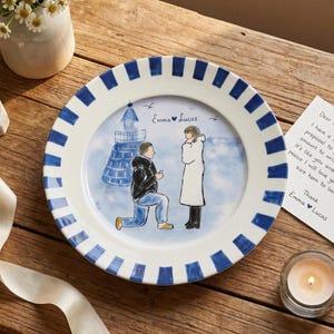 May include: A white ceramic plate with a blue and white striped border. The center features a hand-painted scene of a proposal with a lighthouse in the background. The text "Emma Lucas" is above the couple. A candle and a note are also visible.