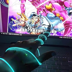 Op de afbeelding: Een hand met gloeiende blauwe neonlichtringen en stervormige accenten, gepositioneerd boven een laptoptoetsenbord. Het scherm toont een kleurrijke illustratie in anime-stijl met Japanse tekst. De scène is verlicht met een koele, donkere toon.