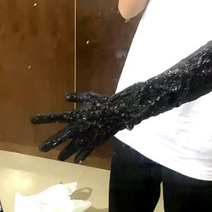 May include: A black, textured arm sleeve with a hand, possibly a costume piece. The sleeve is covered in a dark, possibly wet, material. The background includes a white shirt and a brown wall.