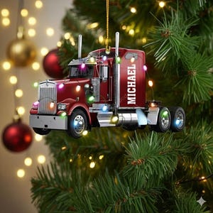 Könnte beinhalten: Ein bordeauxfarbener Weihnachtsbaumschmuck in Form eines Sattelschleppers mit dem Namen "MICHAEL" in weißen Buchstaben auf der Seite. Der LKW ist mit bunten Lichterketten geschmückt und hängt an einer goldenen Schnur. Der Schmuck wird vor einem Weihnachtsbaum präsentiert.