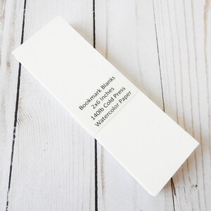 May include: A stack of white bookmark blanks made of 140lb cold press watercolor paper. The blanks are 2x6 inches.
