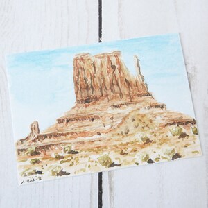 May include: Watercolor painting of a red rock formation in a desert landscape. The sky is a light blue with white clouds. The rock formation is a butte with a flat top and steep sides. The painting is signed "J. Austin '18" in the lower left corner.