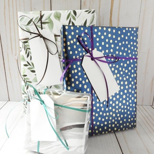 May include: Three gift boxes wrapped in different patterns. The first box is wrapped in white paper with a green leafy design. The second box is wrapped in blue paper with white polka dots. The third box is wrapped in silver paper with a white marbled design. Each box has a white gift tag tied with a ribbon.