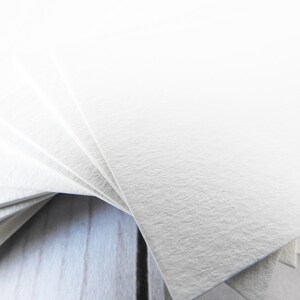 May include: A stack of white textured paper cards fanned out on a light wood surface.