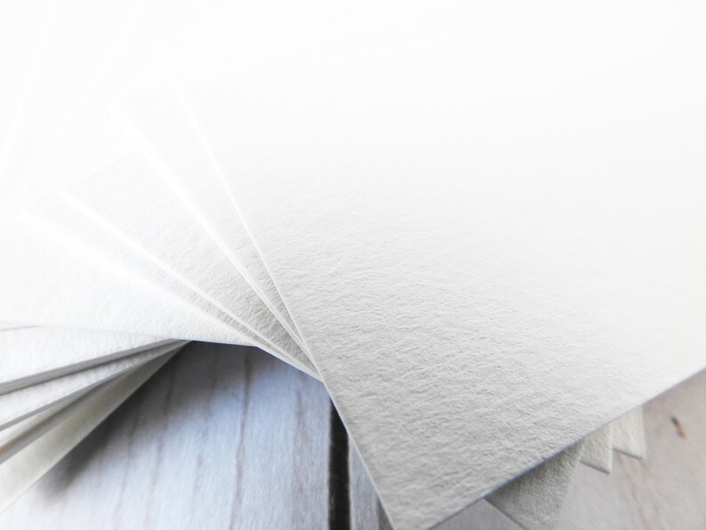 May include: A stack of white textured paper cards fanned out on a light wood surface.
