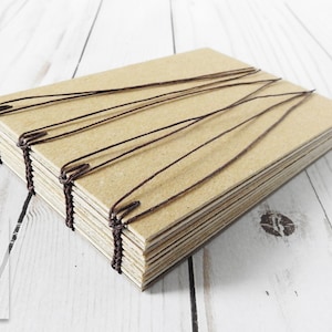 May include: A brown cardboard journal with brown string binding. The journal is made of multiple layers of cardboard and is bound together with brown string. The journal is lying on a white wooden surface.
