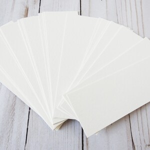 May include: A stack of 20 blank white bookmark cards, fanned out in a semi-circle on a light wood surface.