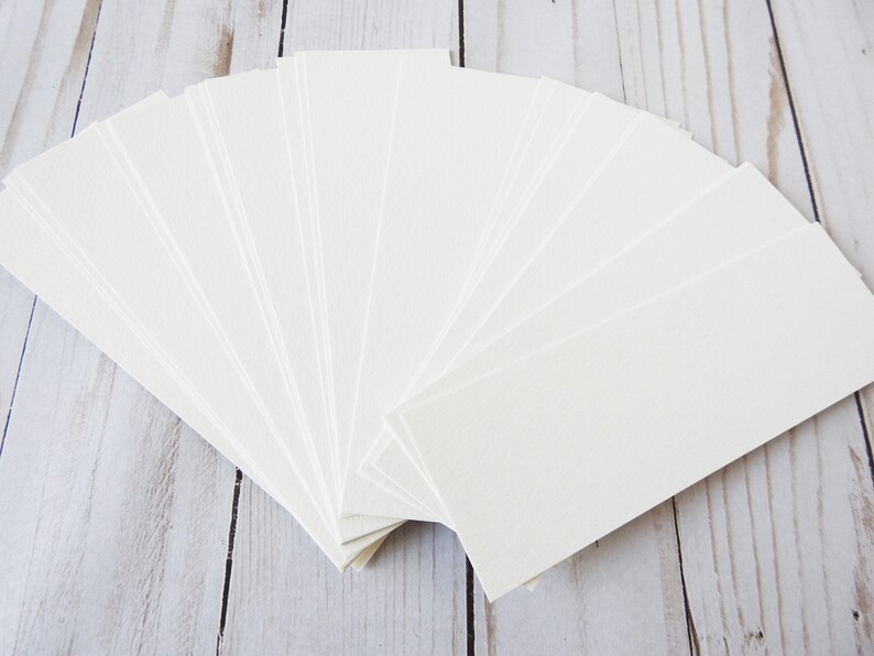 May include: A stack of 20 blank white bookmark cards, fanned out in a semi-circle on a light wood surface.