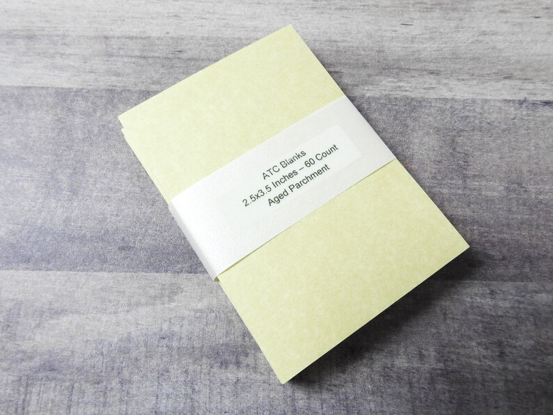 May include: A stack of 60 blank ATC cards in a pale yellow color. The cards are 2.5 x 3.5 inches and are made of aged parchment paper. The cards are wrapped in a white band with the text "ATC Blanks 2.5x3.5 Inches - 60 Count Aged Parchment".
