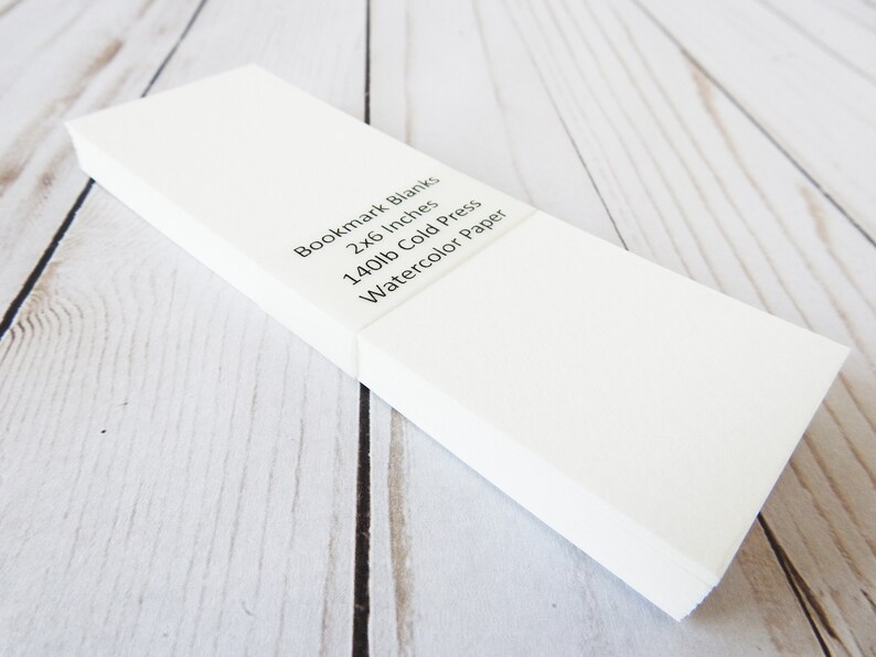May include: A stack of white bookmark blanks made of 140lb cold press watercolor paper. The blanks are 2x6 inches.