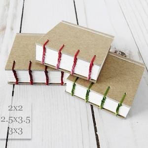 May include: Three small, handmade journals with brown paper covers and white pages. The journals are bound with red, pink, and green thread. The text "2x2", "2.5x3.5", and "3x3" is written on a white surface below the journals.