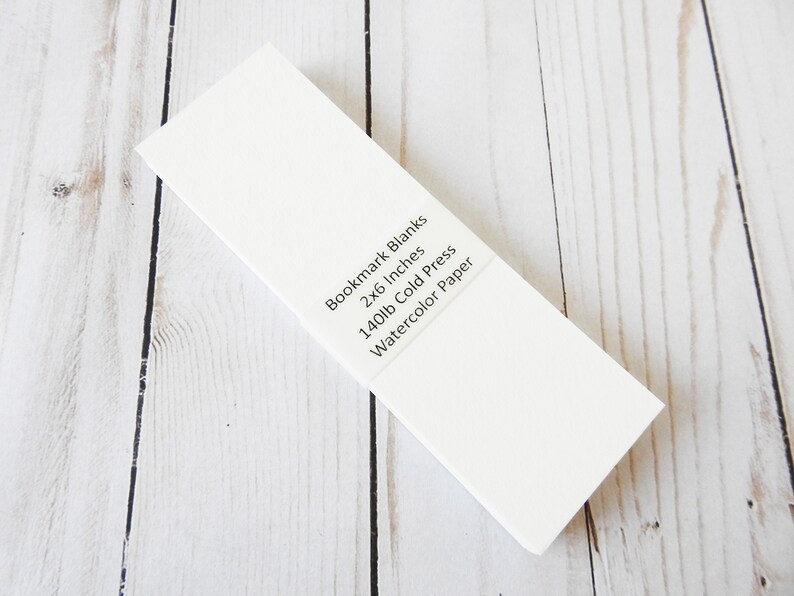 May include: A stack of white bookmark blanks made of 140lb cold press watercolor paper. The blanks are 2x6 inches.