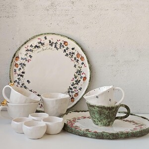 Può includere: Una collezione di stoviglie in ceramica fatte a mano, tra cui piatti, tazze e piccole ciotole. I pezzi bianchi presentano una superficie strutturata e sono adornati da disegni floreali e di bacche nei toni del verde, blu e arancione. Alcune tazze hanno dettagli dorati.