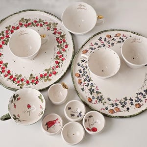 Op de afbeelding: Een verzameling wit keramisch serviesgoed met handgeschilderde fruit- en bloemmotieven. De set bevat grote borden, theekopjes en kleine kommen, met gouden accenten op sommige handvatten. De borden hebben aardbei- en bosbessenmotieven.