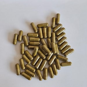 Könnte beinhalten: Eine Ansammlung von zahlreichen, zylindrischen, goldbraunen Kapseln auf einer weißen Oberfläche. Die Kapseln haben ein glattes, metallisches Aussehen, was auf ein Nahrungsergänzungsmittel oder Medikament hindeutet. Die Kapseln sind von einheitlicher Größe und Form.