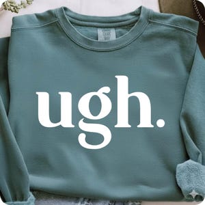 May include: A teal crewneck sweatshirt featuring the word "ugh." printed in large, bold white serif letters across the center. The garment has a ribbed collar and long sleeves, with a visible brand label inside the neckline.