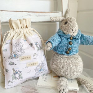 May include: A small, white canvas bag with a drawstring closure and a Peter Rabbit print. The bag has the words "bunny kisses" printed on it. A knitted grey bunny wearing a blue sweater is sitting on a book.