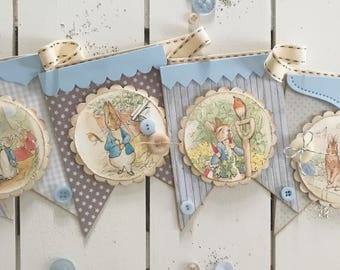 May include: A blue and brown banner with a scalloped edge features illustrations of Peter Rabbit in various scenes. The banner is decorated with blue and white buttons and a white ribbon.