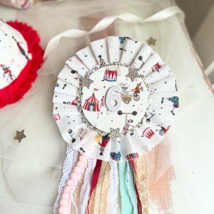 May include: A white and red circus-themed rosette with a number 6 in the center. The rosette has a pleated design with a glittery border and star accents. Colorful ribbons and lace hang from the bottom, creating a festive look.