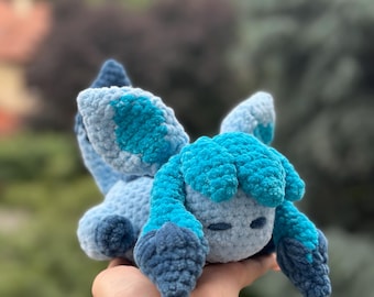 Puede incluir: Un peluche hecho a mano en tonos azules, con un diseño de animal estilizado. El peluche tiene un aspecto suave y texturizado, con detalles como orejas y una cola. El juguete se sostiene en una mano sobre un fondo verde borroso.