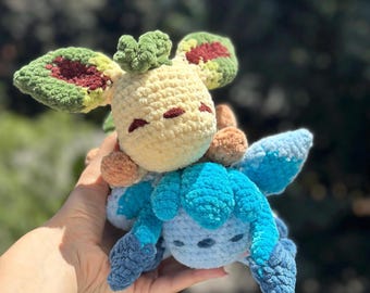 Puede incluir: Dos peluches de ganchillo. El superior es amarillo con detalles verdes y granates, y el inferior es azul. Los juguetes tienen orejas grandes y se sostienen en una mano sobre un fondo verde borroso.