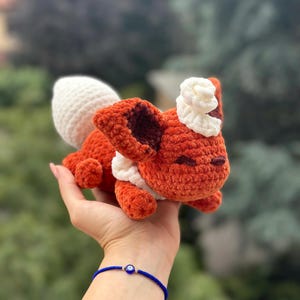 Puede incluir: Un peluche de zorro de ganchillo naranja y blanco hecho a mano. El zorro tiene una cola blanca, detalles blancos alrededor del cuello y la cabeza, y orejas internas de color marrón oscuro. El peluche se sostiene en una mano sobre un fondo verde borroso.