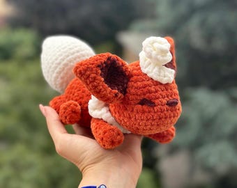 Puede incluir: Un peluche de zorro de ganchillo naranja y blanco hecho a mano. El zorro tiene una cola blanca, detalles blancos alrededor del cuello y la cabeza, y orejas internas de color marrón oscuro. El peluche se sostiene en una mano sobre un fondo verde borroso.