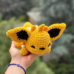 May include: A handmade, yellow crochet plush toy with black accents on the wings and face. The plush has a soft, textured appearance and is held in a hand. The background is a blurred green and brown.