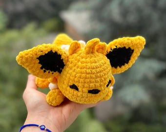Puede incluir: Un peluche amarillo hecho a mano con ganchillo, con detalles negros en las alas y la cara. El peluche tiene un aspecto suave y texturizado y se sostiene en una mano. El fondo es un desenfoque verde y marrón.