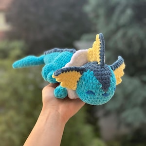 Peut inclure: Une peluche Vaporeon faite à la main au crochet. La peluche est principalement turquoise, avec des détails jaunes, blancs et bleu marine. Elle a un visage souriant et est tenue dans une main. C'est un jouet câlin.